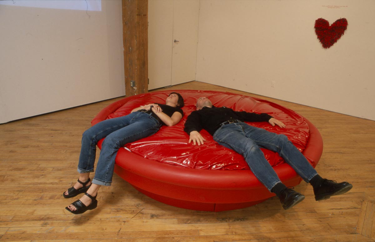 Clara Ursitti: Installation view, man and woman laying on circular sculpture.