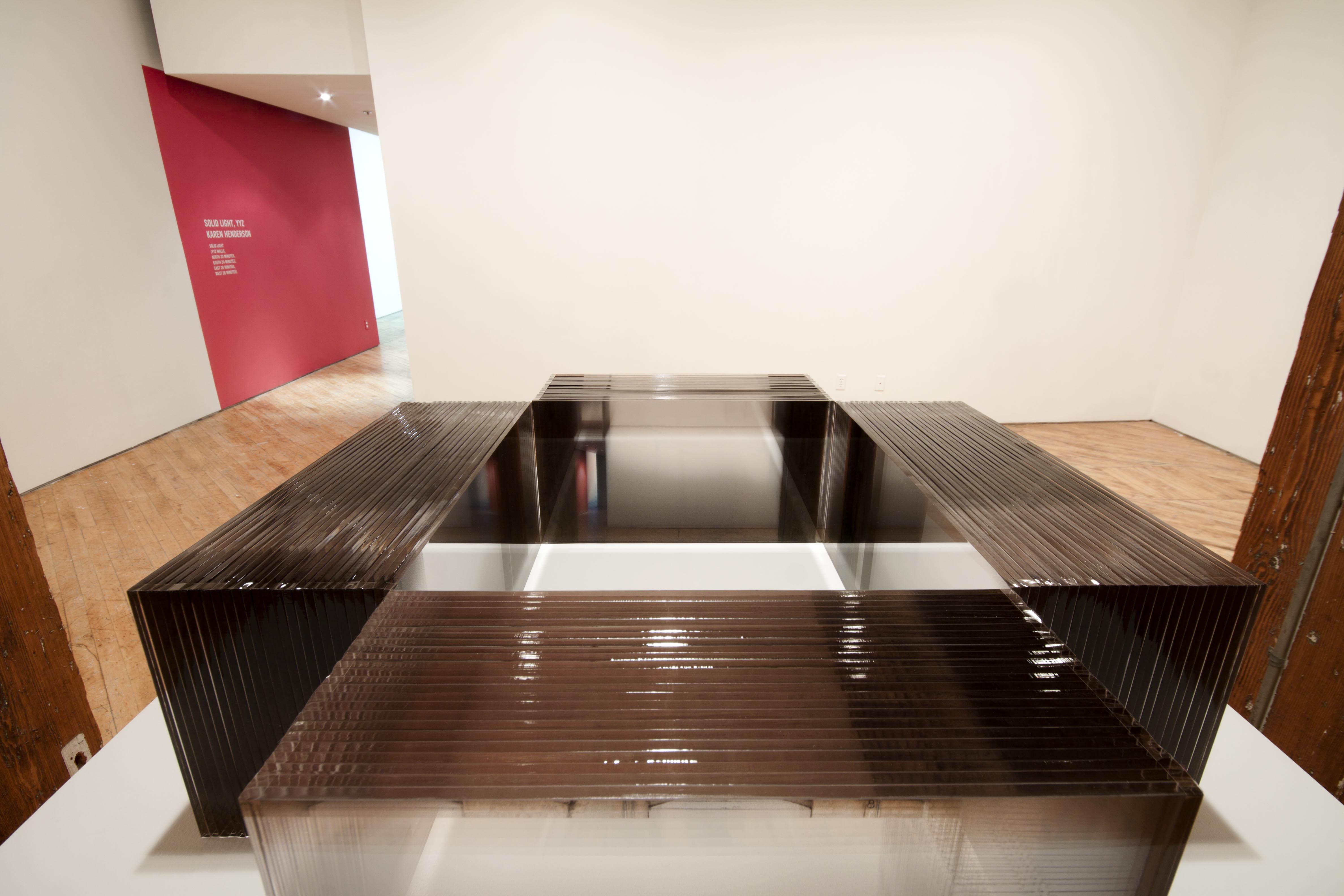 Karen Henderson, plinth, on top of which 4 acrylic rectangular blocks are placed so that each of their inside corners are touching. Each one is made up of 26 sheets of acrylic fused together. Each sheet has a photographic panorama printed on it a composite image of the gallery wall