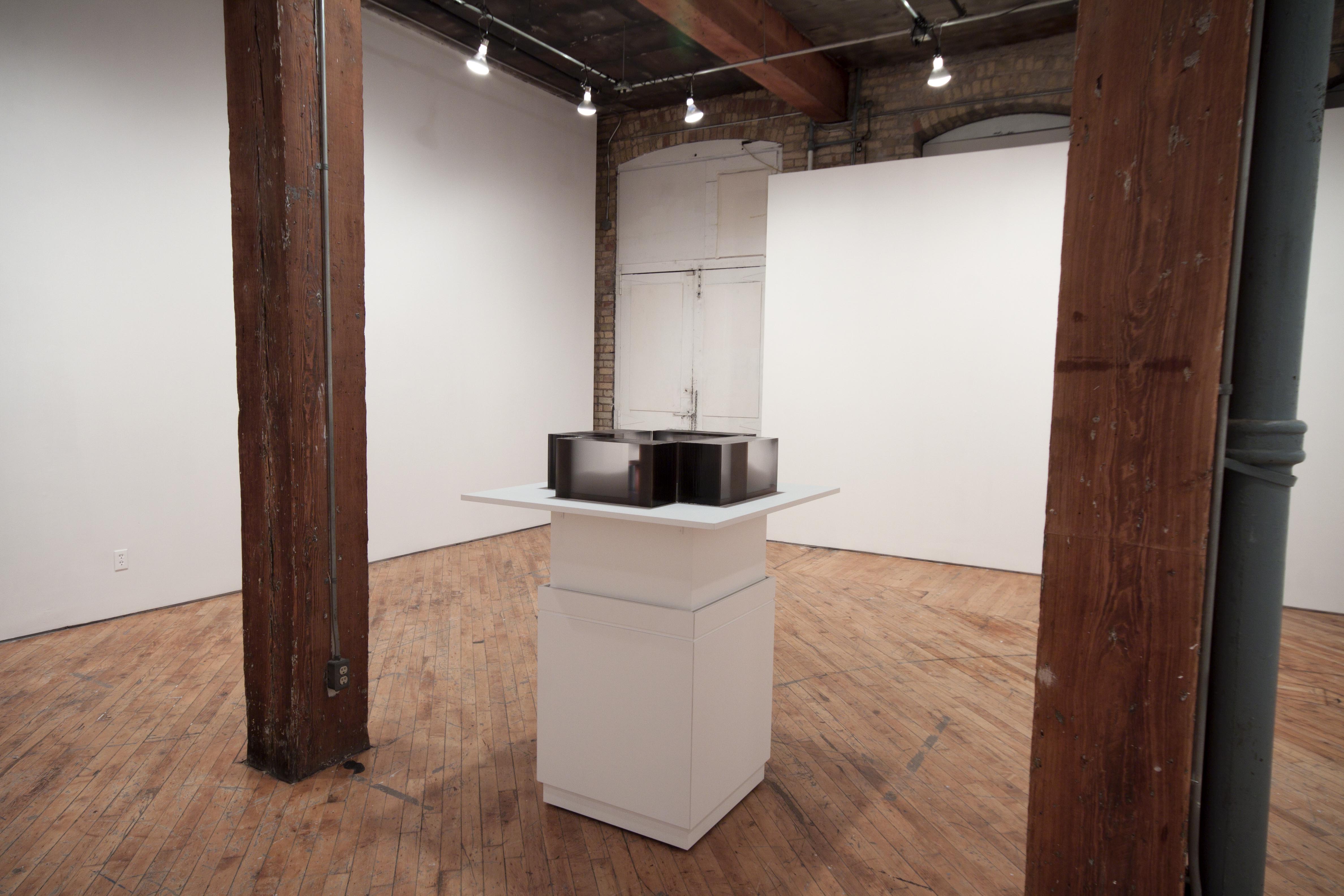 Karen Henderson, plinth, on top of which 4 acrylic rectangular blocks are placed so that each of their inside corners are touching. Each one is made up of 26 sheets of acrylic fused together. Each sheet has a photographic panorama printed on it a composite image of the gallery wall