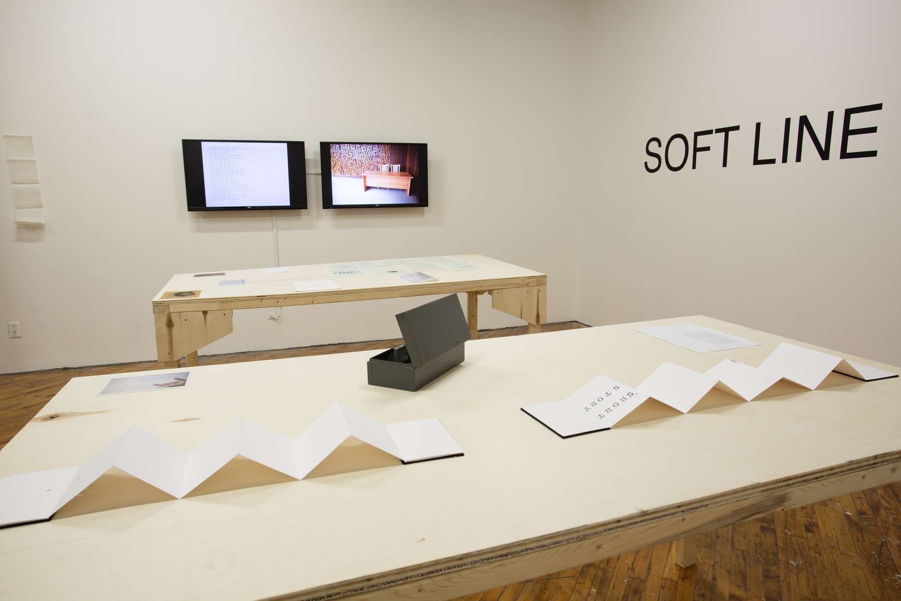 Yusuf Hassan, installation view, two tables arranged side by side, both contain printed material, and objects, two monitors in the background on the wall.