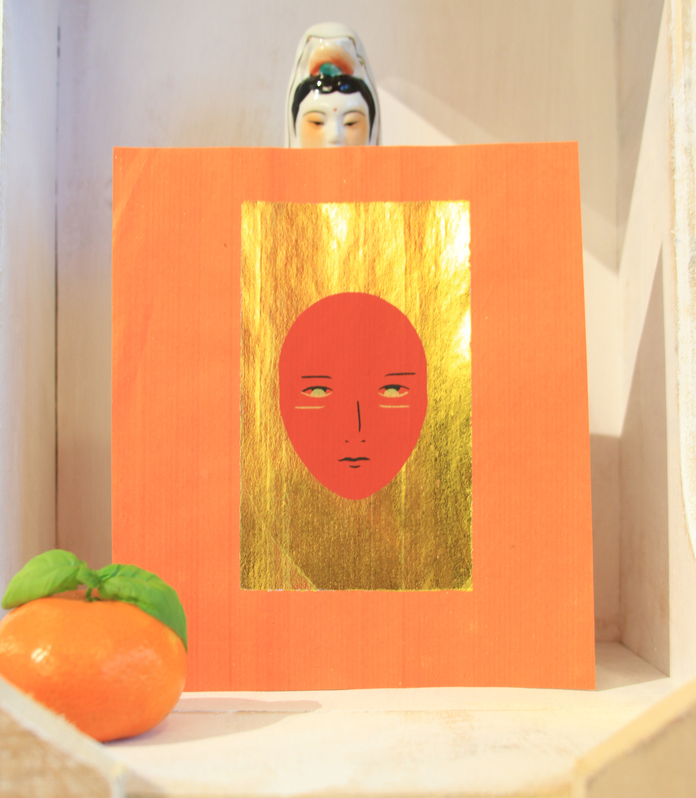 An orange card rests against a human figurine. the card has a gold rectangle with a face on it. A mandarin fruit sits in front of the card.