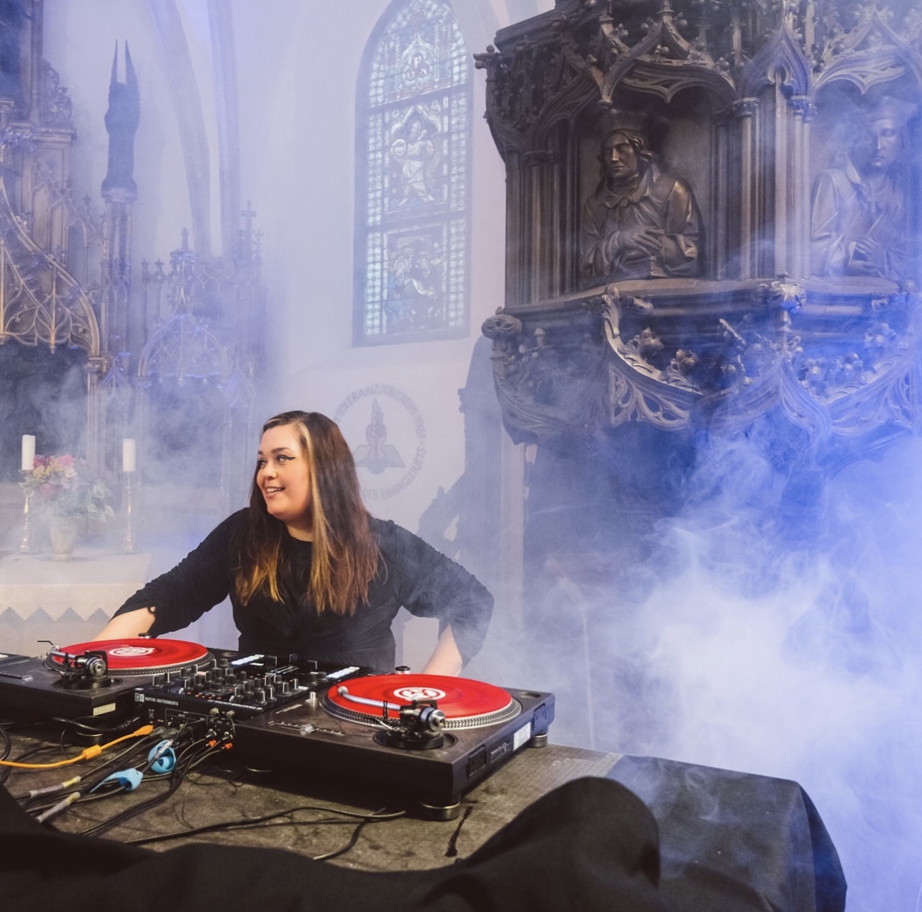 Woman stands behind DJ booth, she is enveloped in smoke